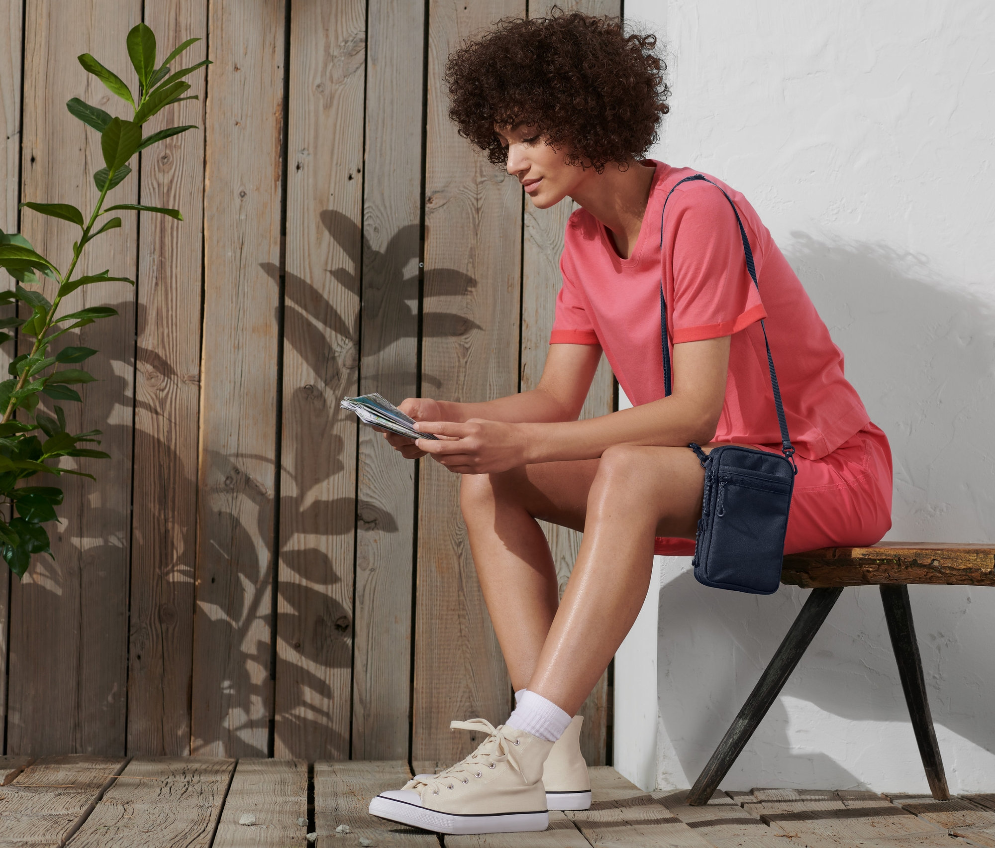 Eine Frau sitzt auf einer Bank und liest. Sie trägt ein rosa T-Shirt und Shorts und eine Umhängetasche über der Schulter.