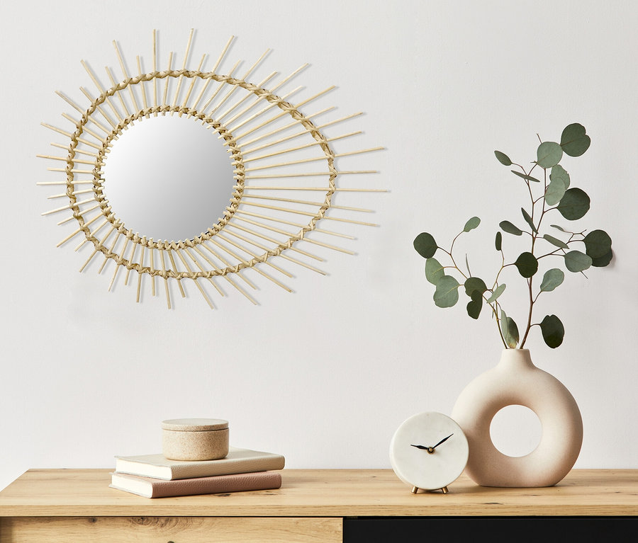 An einer weißen Wand hängt ein Kayoom Rattan-Spiegel »Aina« 125. Darunter befinden sich auf einer Kommode eine Vase mit einer Pflanze, eine Uhr und Bücher.