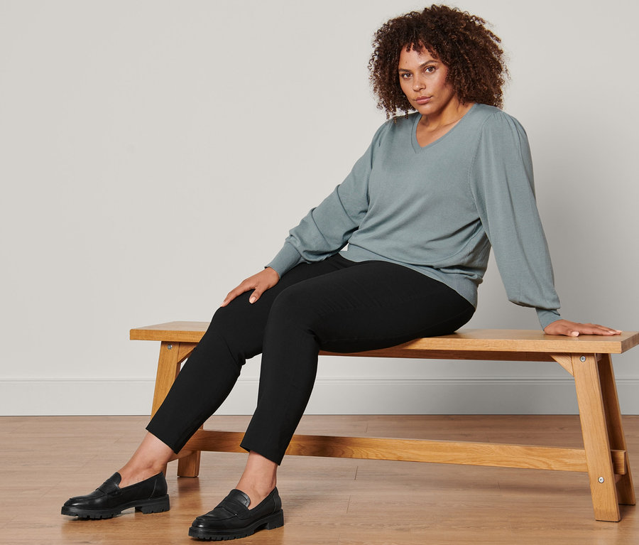 Frau sitzt auf einer Bank und trägt einen Feinstrickpullover, eine Stretchhose und Leder-Loafer.
