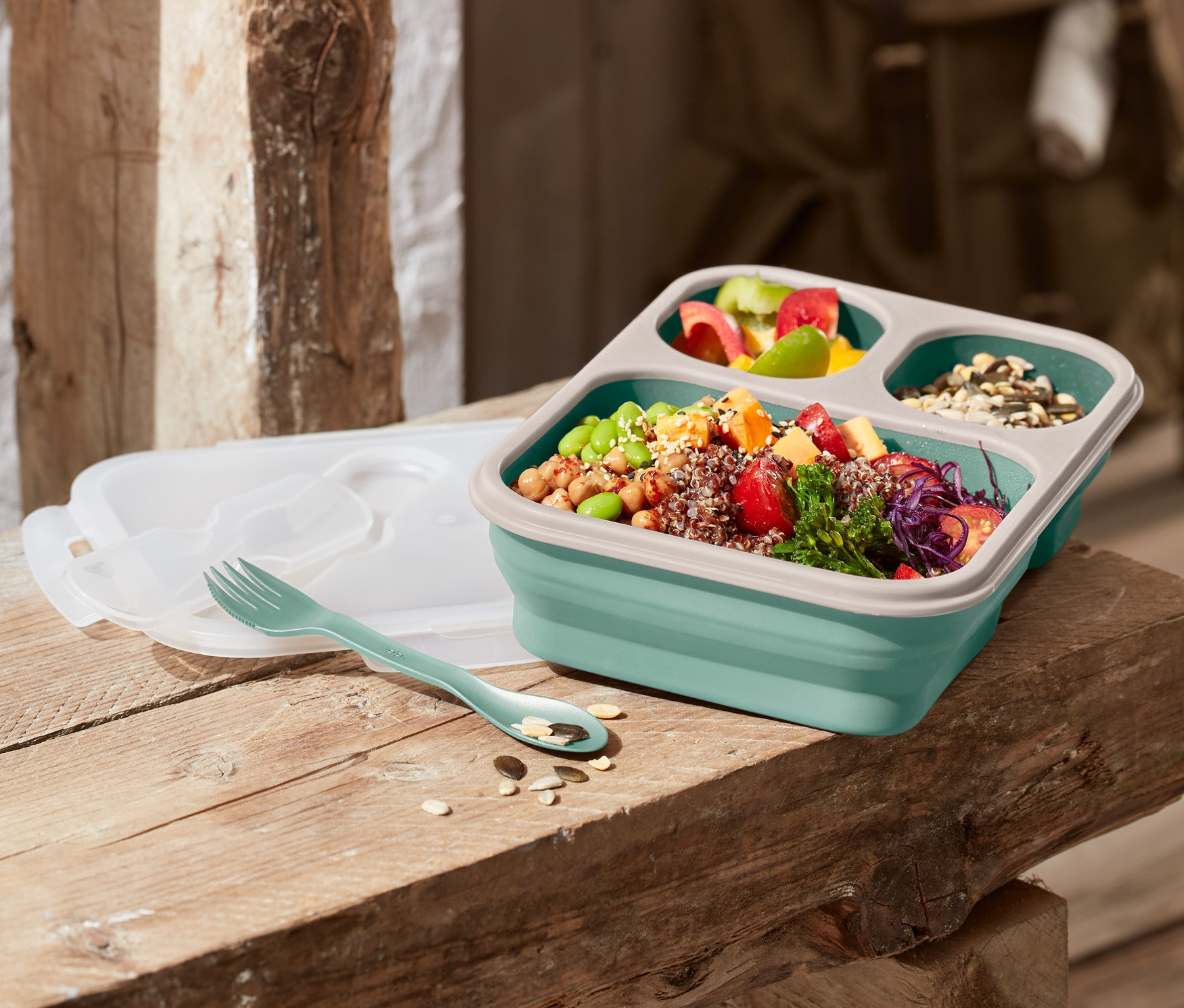 Eine faltbare Lunchbox steht auf einer Holzbank, daneben liegen eine Gabel und ein Deckel.