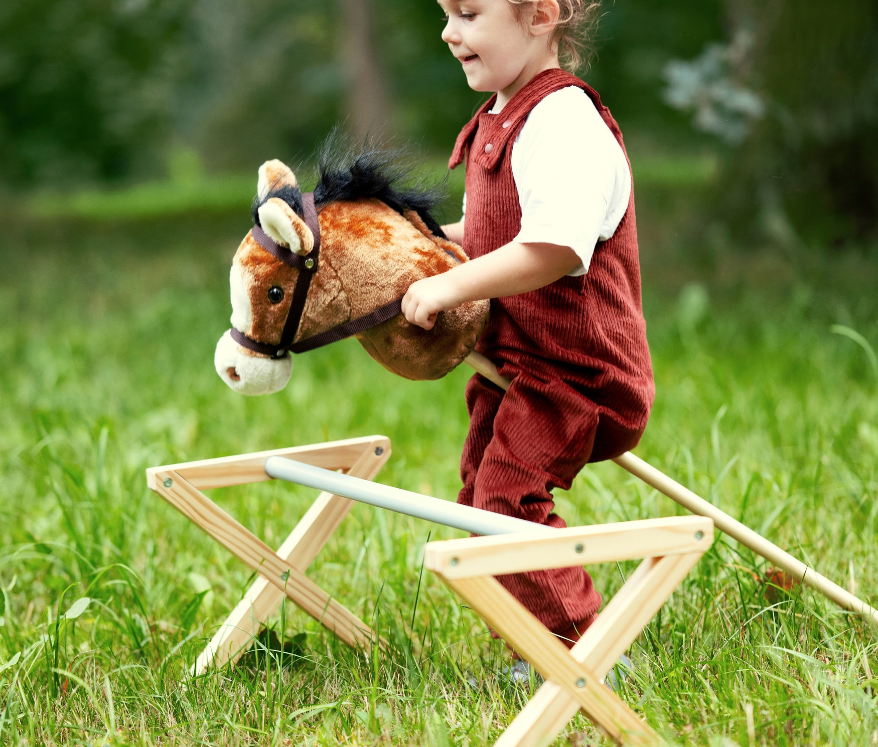 Mädchen in rotem Overall reitet auf einem braunen small foot Steckenpferd »Nico« auf einer Wiese.