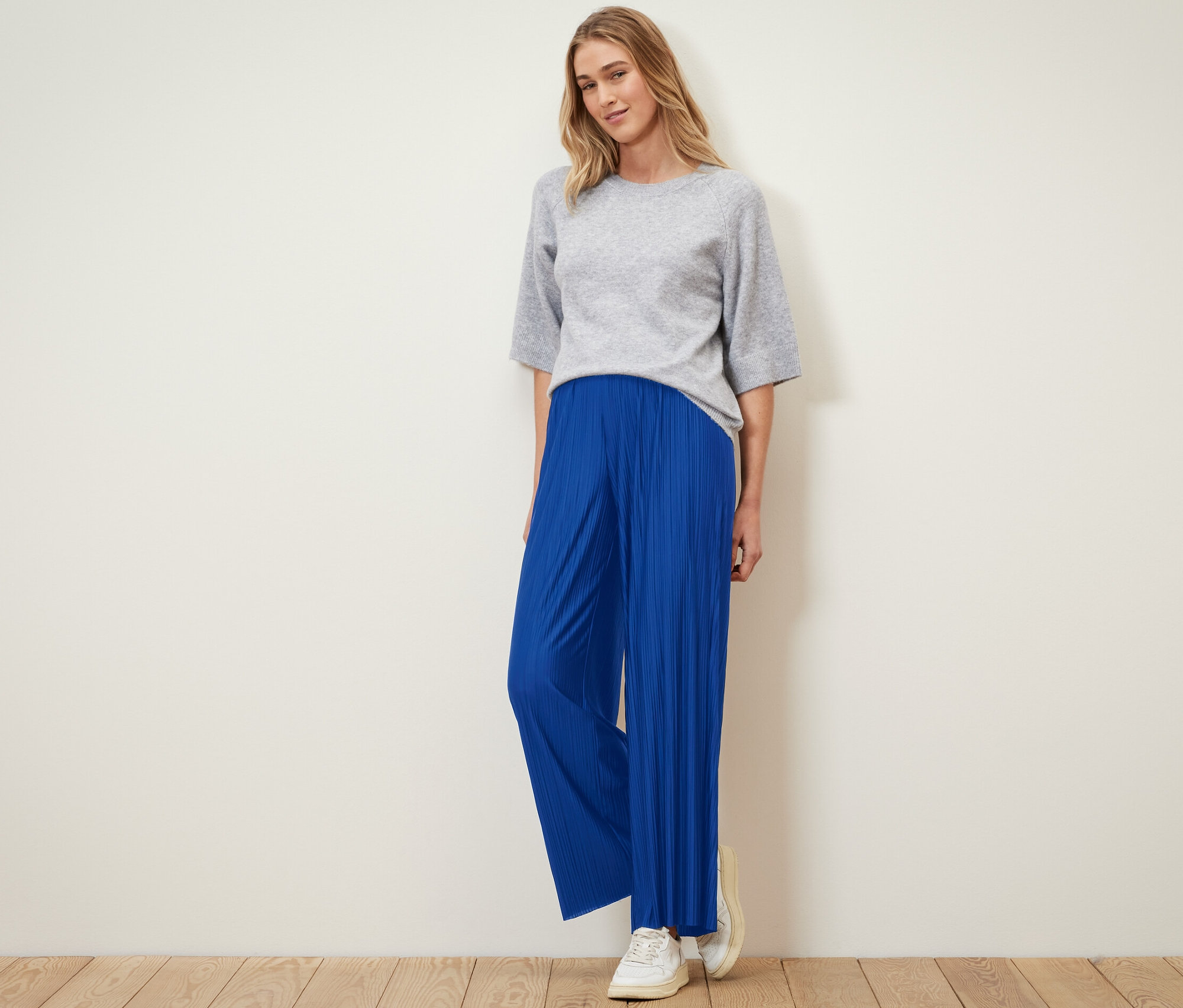 Eine Frau steht und trägt eine kobaltblaue Plisseehose und ein graues Oberteil.
