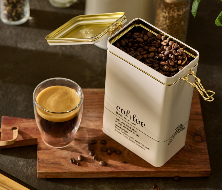 Eine geöffnete Kaffeedose, coffee dictionary, gefüllt mit Kaffeebohnen, steht neben einem Glas Kaffee auf einem Holzbrett.