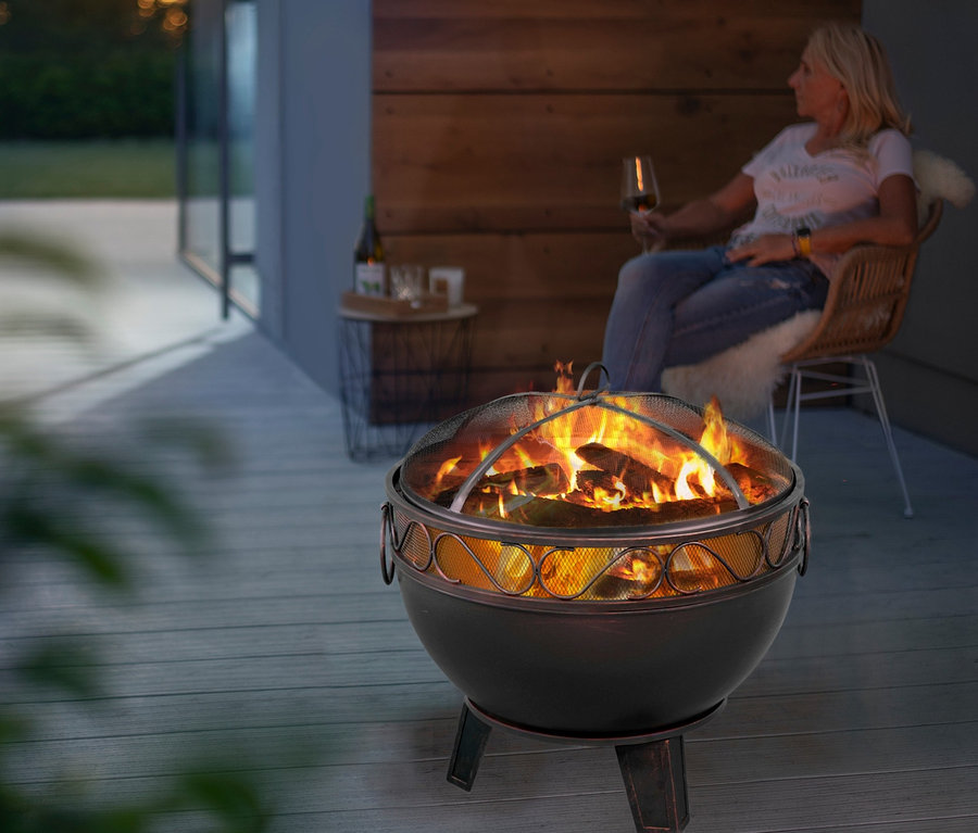 Frau sitzt draußen mit einem Getränk in der Nähe einer Buschbeck-Feuerschale mit Funkenschutzhaube und Feuerrost.
