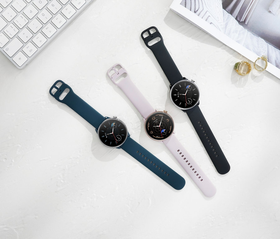 Drei Amazfit GTR Mini Smartwatches (blau, pink und schwarz) liegen auf einer weißen Oberfläche neben einer Tastatur und einer Zeitschrift.