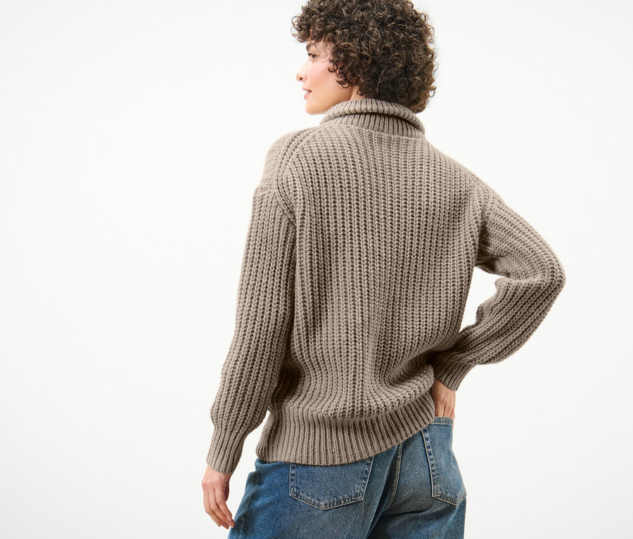 Frau posiert mit dem Rücken zur Kamera und trägt einen taupefarbenen Grobstrick-Troyer und blaue Jeans.