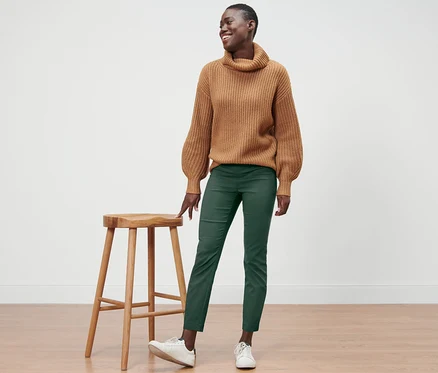 Eine lächelnde Frau steht neben einem Holzhocker und trägt einen braunen Pullover und eine grüne Stretchhose.