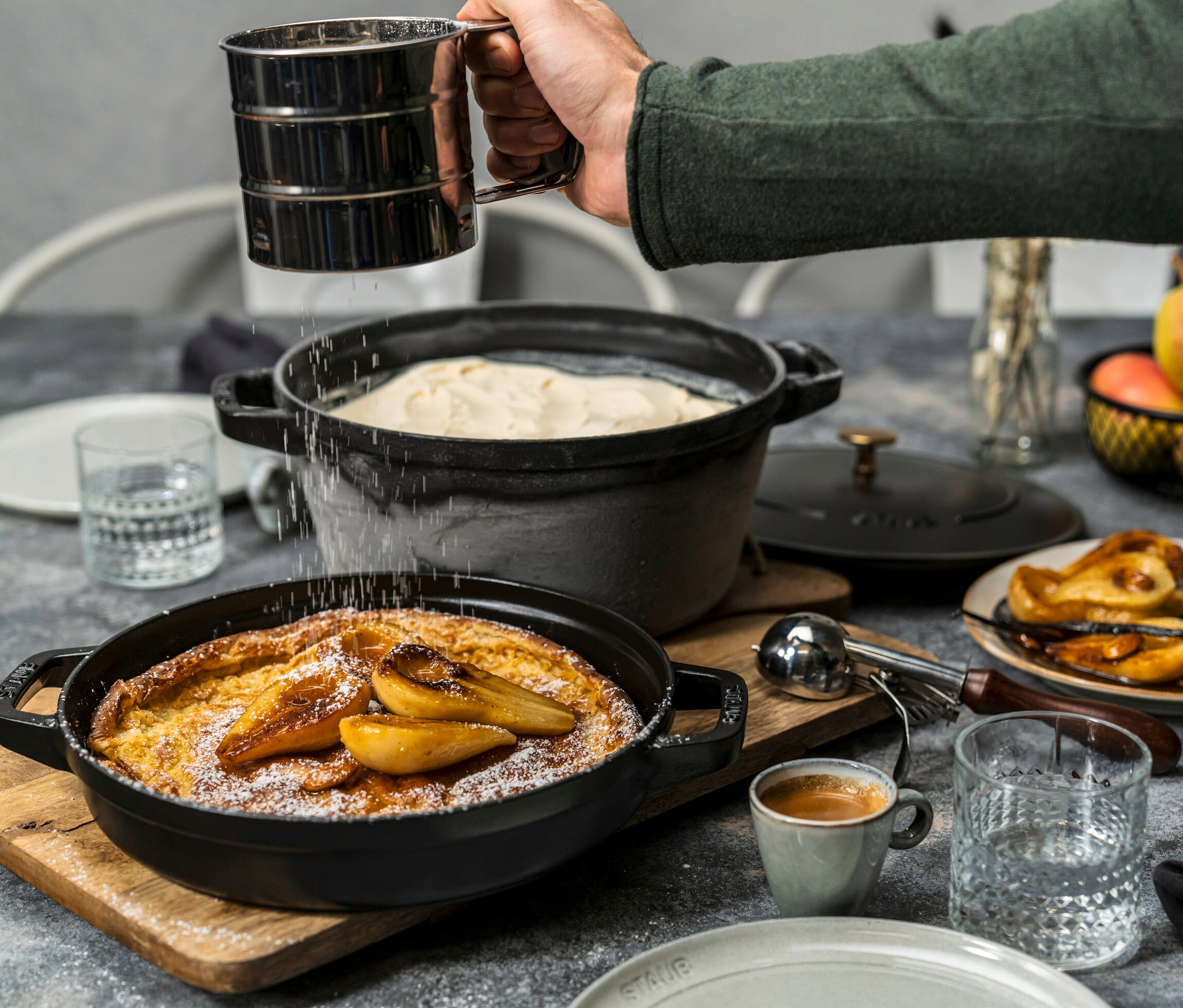 Eine Hand streut Puderzucker auf einen Dutch Baby mit Birnen; daneben ein Staub Gusseisen-Kochtopf-Set mit Teig.