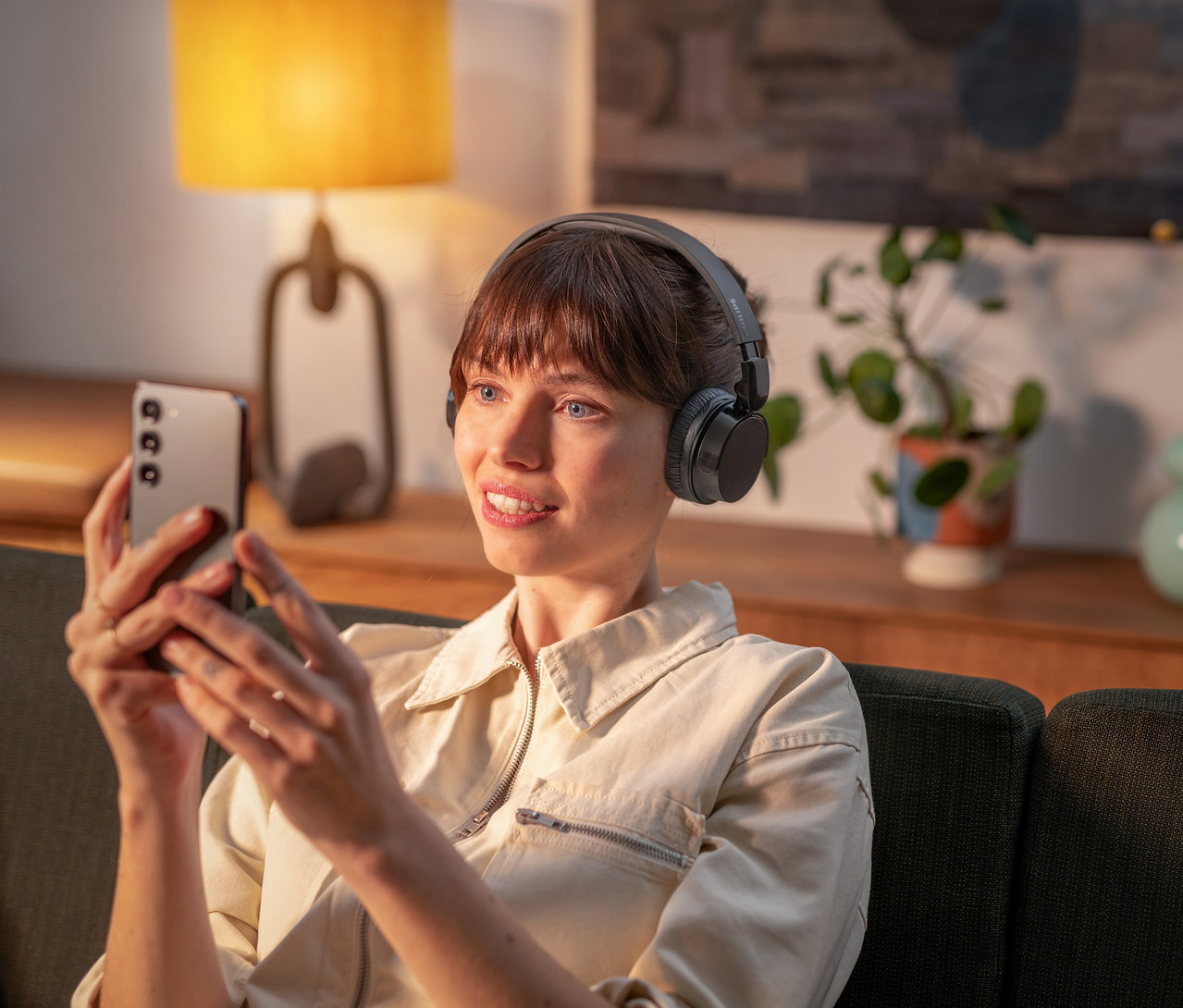 Frau mit Philips Bluetooth®-Kopfhörern »H3209« schaut auf ein Handy, während sie auf einem Sofa sitzt.