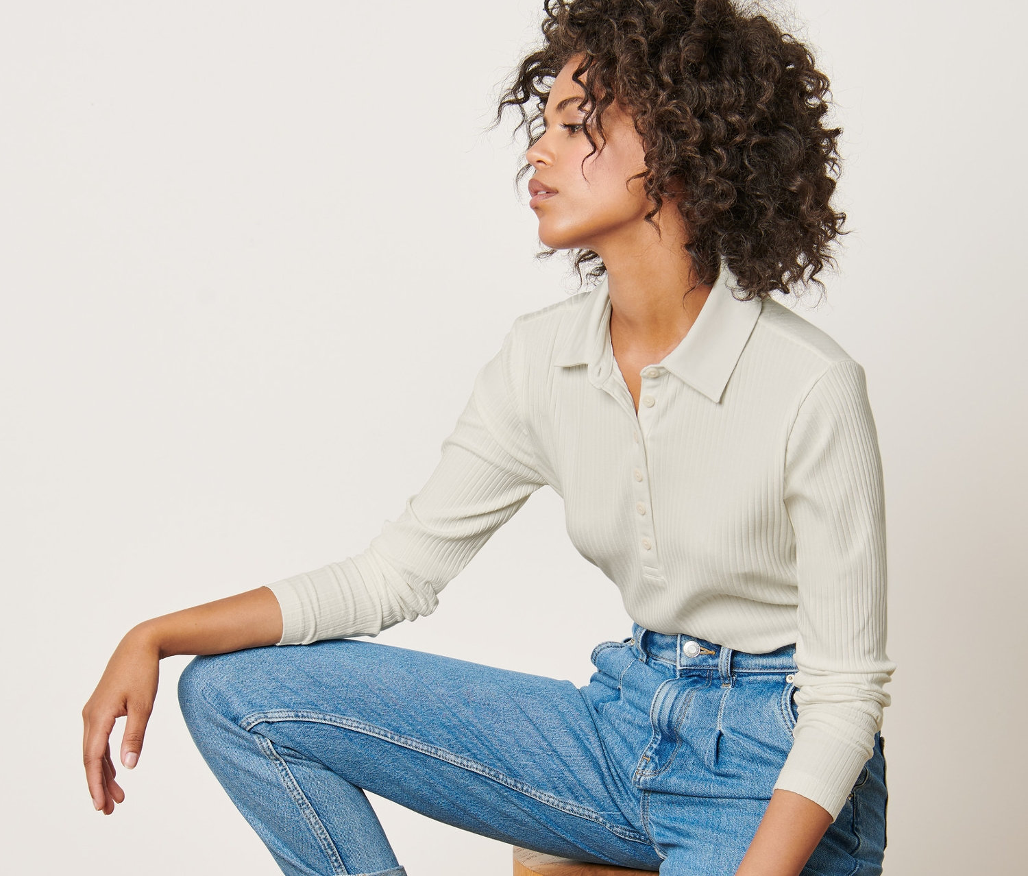 Frau sitzt auf einem Holzhocker und trägt ein geripptes Langarmshirt mit Polokragen, Jeans und Turnschuhe.