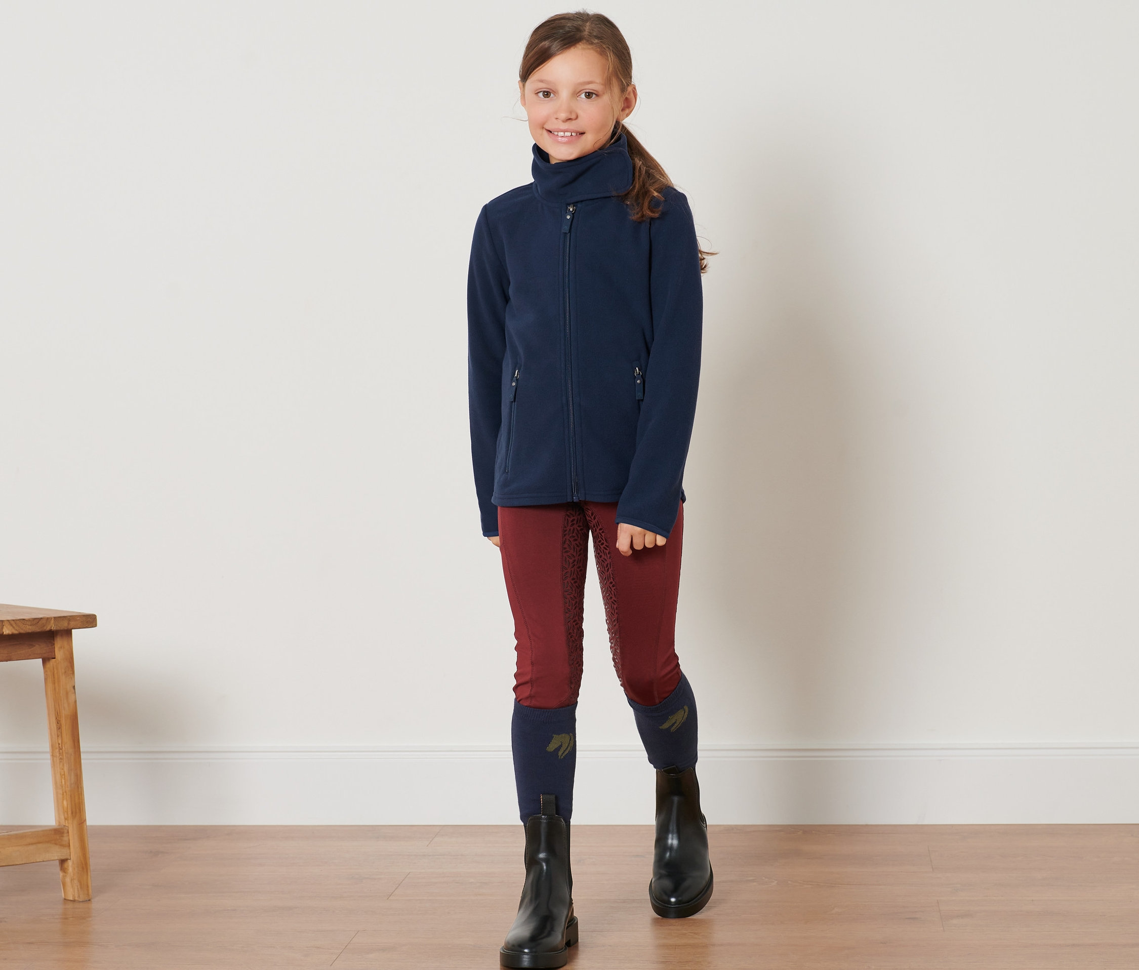 Mädchen steht und trägt eine Kinder-Fleecejacke, Reithosen und schwarze Stiefel.