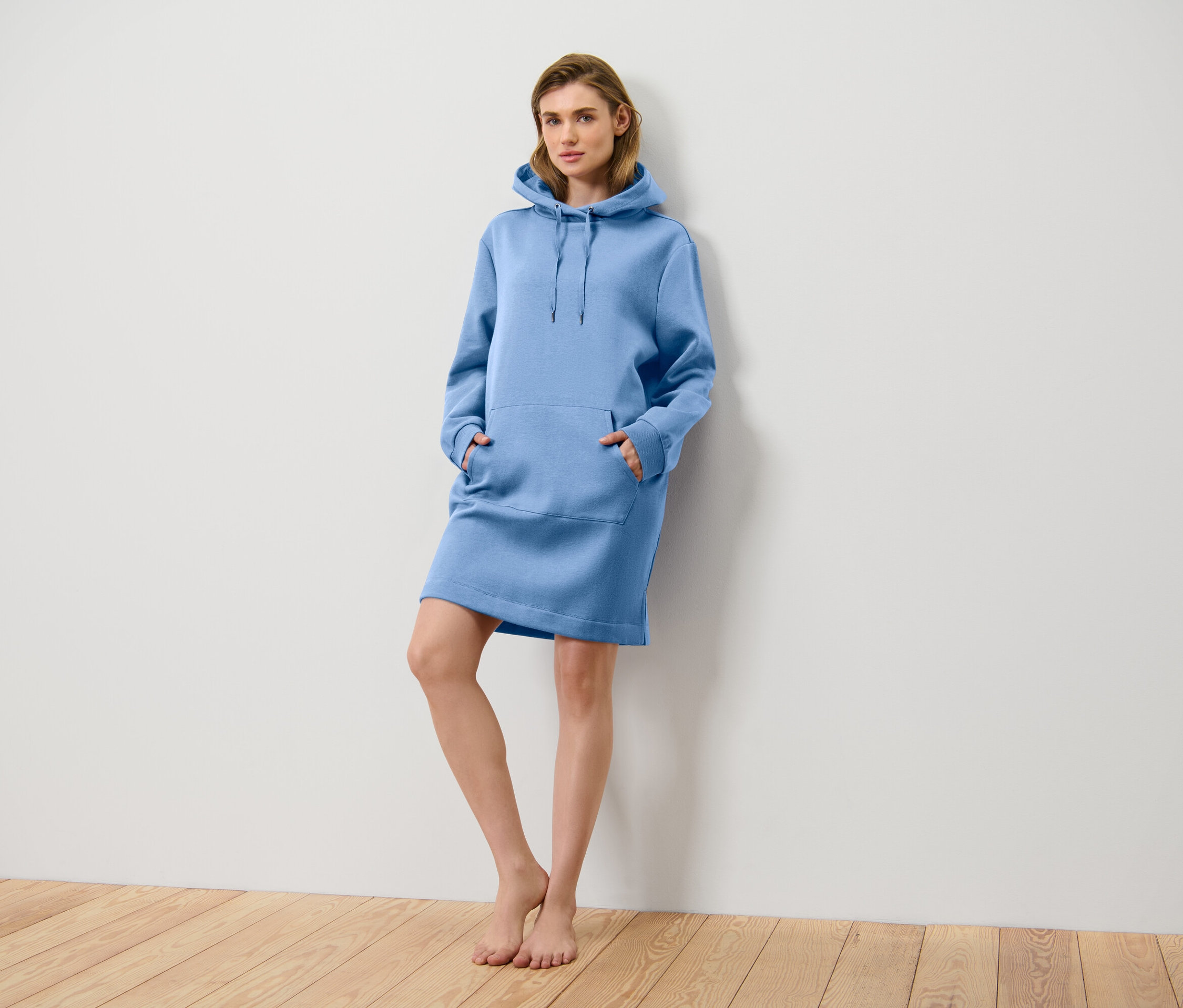 Eine Frau steht an einer Wand und trägt ein blaues Hoodie-Kleid mit Kapuze und Taschen.