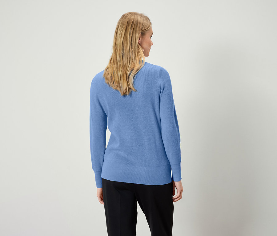 Frau mit blonden Haaren trägt einen hellblauen Pullover und eine schwarze Hose, von hinten gesehen.