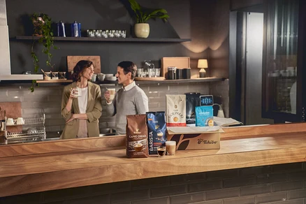 Zwei Personen halten Kaffeetassen in einer Küche hinter Tchibo Kaffeepackungen.