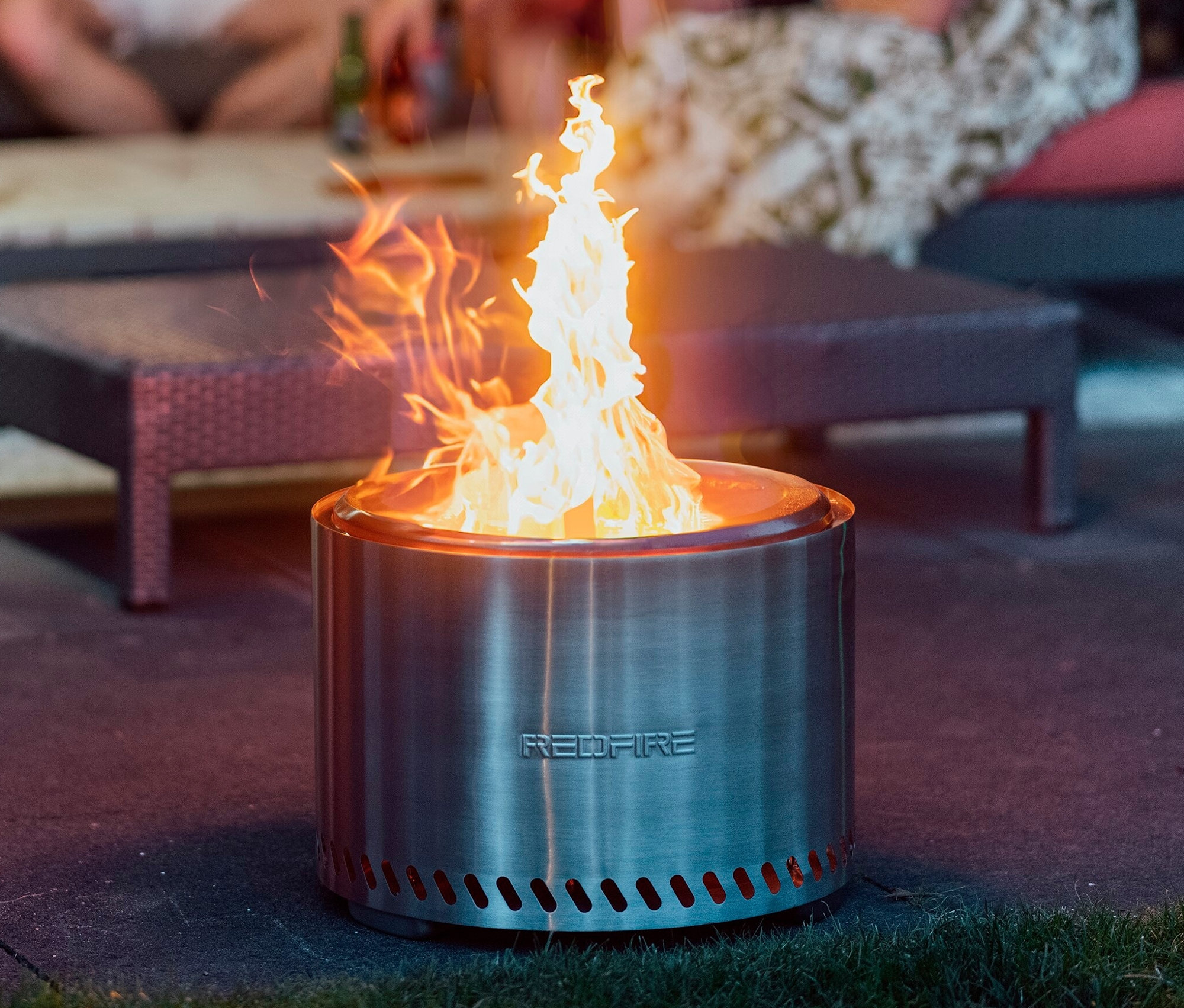 Silberfarbene RedFire Feuerschale »Volcano Extra Large« mit Feuer, im Hintergrund sitzende Personen.