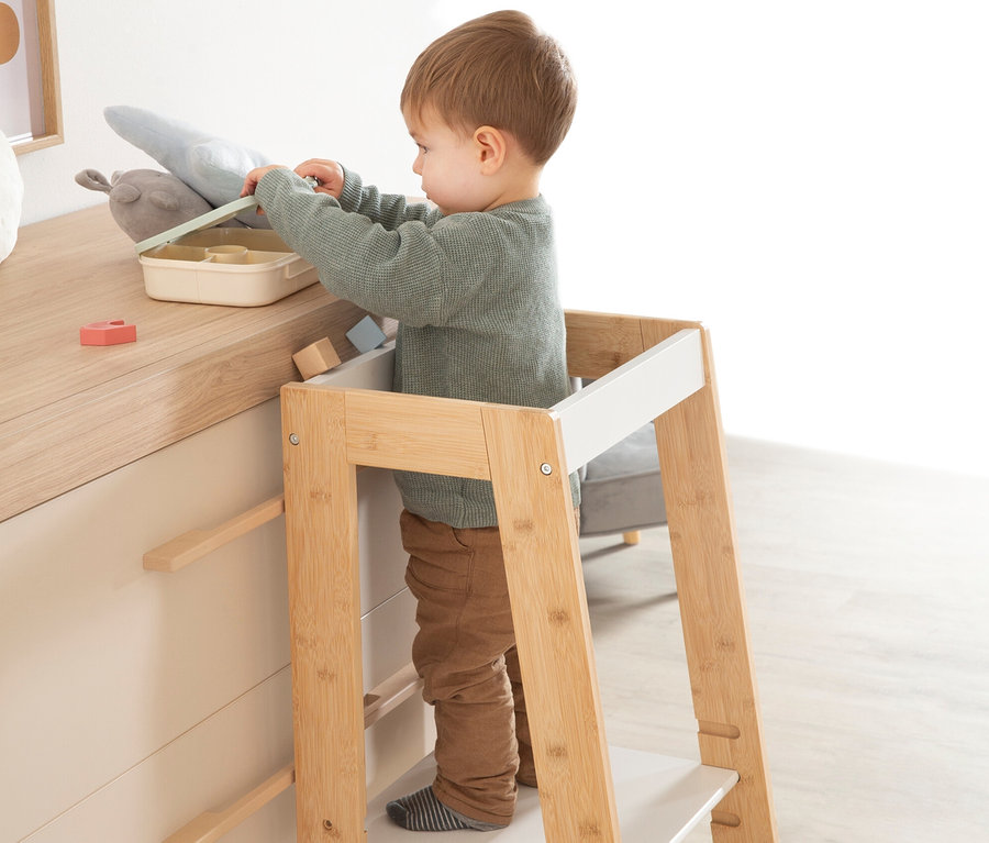 Ein Kind steht auf dem weißen roba Lernturm holz an einem Tisch und spielt mit Spielzeug.