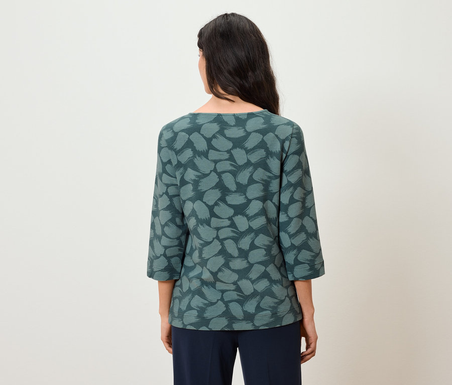 Rückansicht einer Frau mit langen, dunklen Haaren, die ein blaugrünes Oberteil mit Dreiviertelärmeln und eine dunkelblaue Hose trägt.