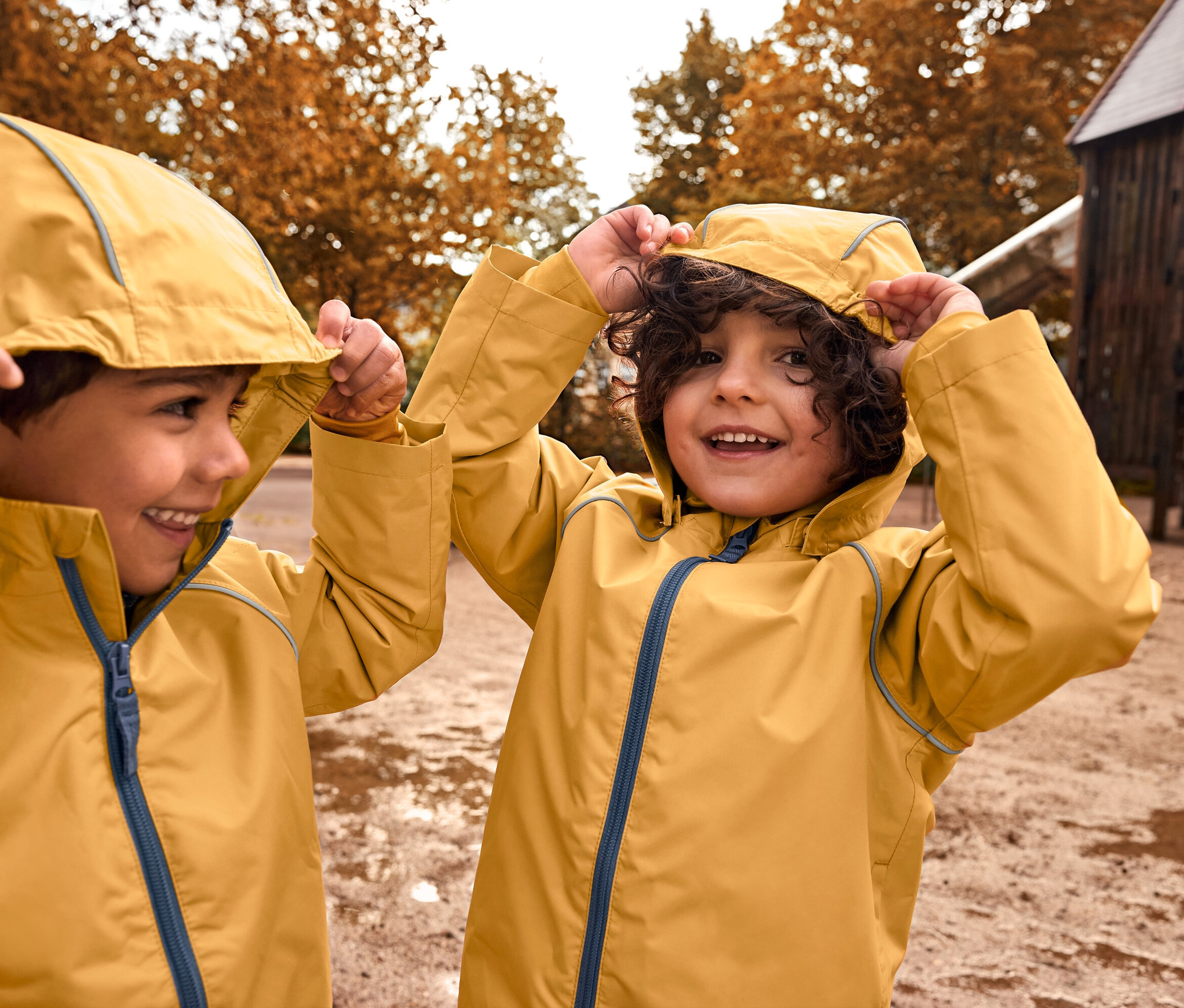 Zwei Jungen in gelben Regenjacken ziehen ihre Kapuzen auf einem Spielplatz hoch.
