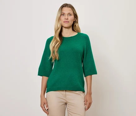 Blonde Frau trägt einen grünen Pullover mit Dreiviertelärmeln und eine beige Hose.
