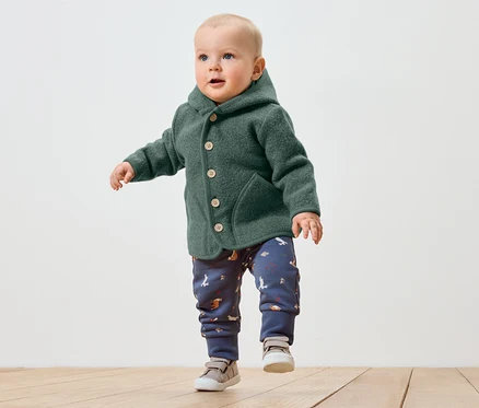Kleinkind in grüner Wolljacke mit Kapuze und gemusterter Hose macht erste Schritte auf Holzboden.