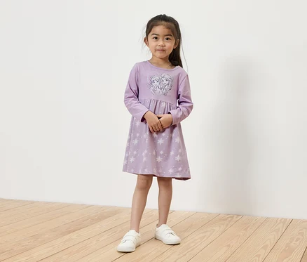 Ein Mädchen steht und trägt ein hellviolettes, langärmliges Kleid mit einem Elsa- und Anna-Motiv und weiße Turnschuhe.