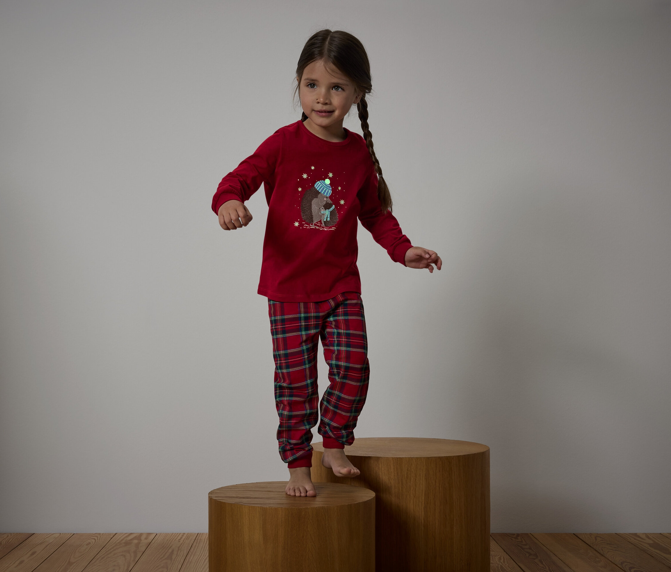 Mädchen im roten Pyjama mit karierter Hose steht auf Holzhockern.