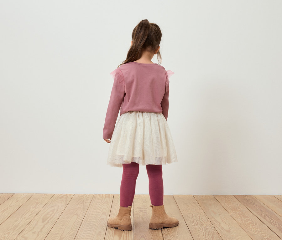 Mädchen mit dem Rücken zur Kamera trägt einen rosa Pullover, einen glitzernden Rock, rosa Leggings und braune Stiefel.