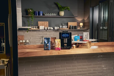 Auf einer Holzplatte stehen eine Kaffeemaschine, drei Kaffeepackungen und eine Kiste mit Kaffee.