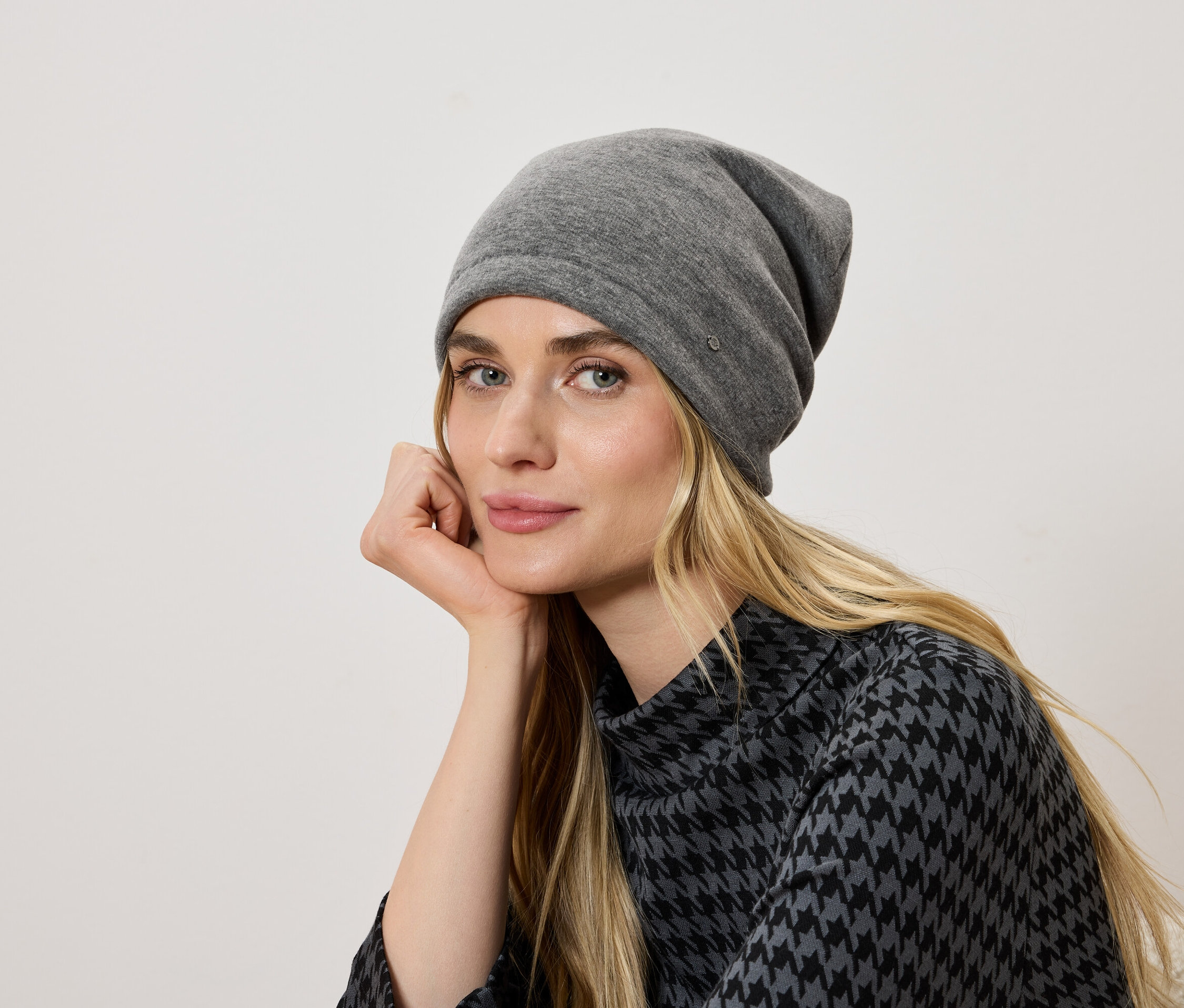 Porträt einer Frau mit blonden Haaren, die eine graue Mütze und eine Bluse mit Hahnentrittmuster trägt.