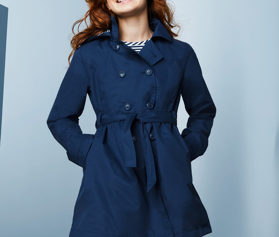 Lächelndes junges Mädchen posiert in einem marineblauen Allwetter-Trenchcoat mit recyceltem Material.