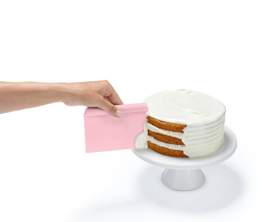 Eine Hand trägt mit einem pinkfarbenen Spachtel Zuckerguss auf eine dreischichtige Torte auf, die auf einem weißen Sockel steht.