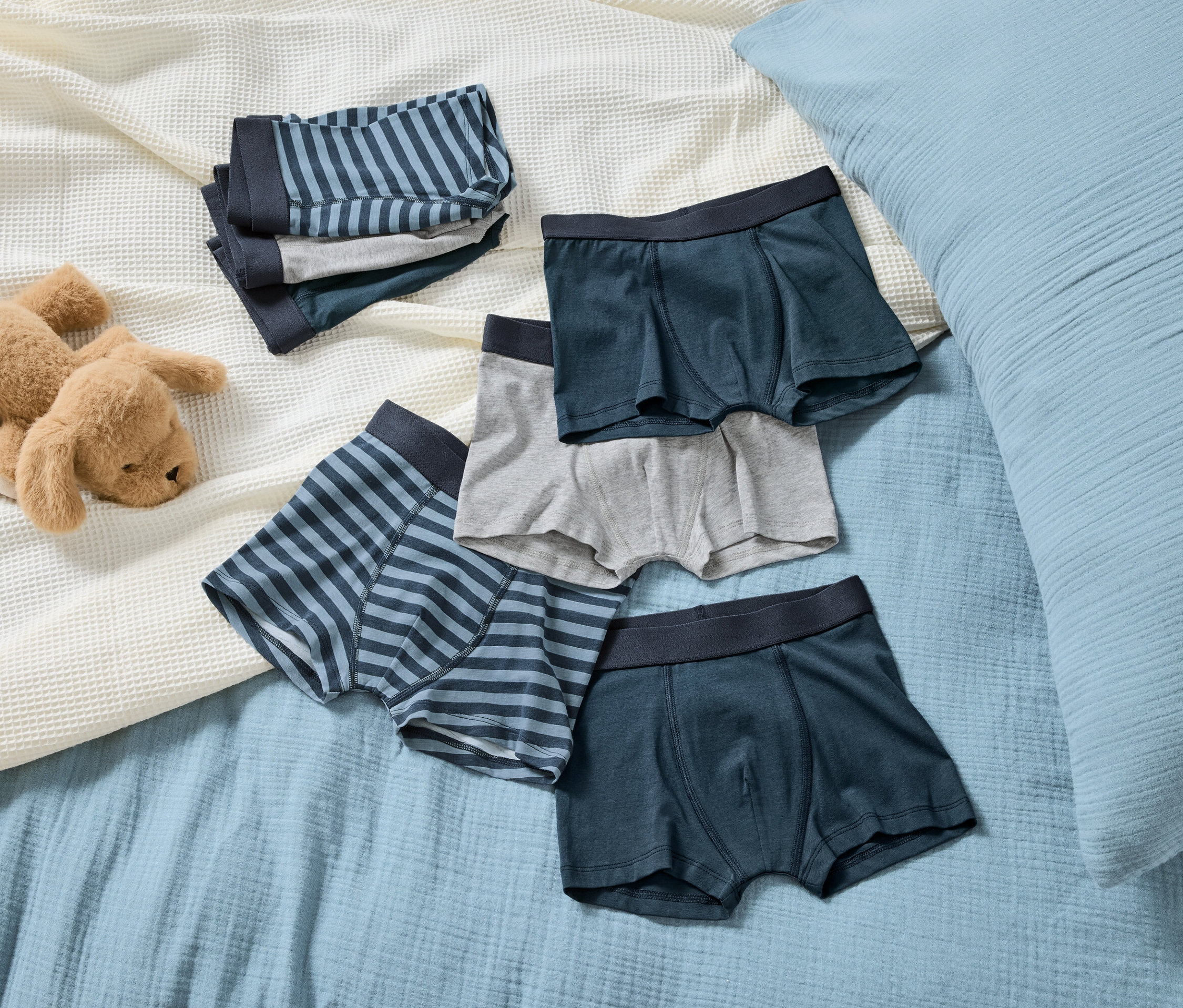 Auf einer hellblauen Decke liegt ein brauner Stoffhund neben einem Stapel gestreifter und unifarbenen Jungen-Boxershorts.