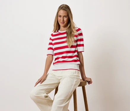 Blonde Frau sitzt auf einem Holzhocker, trägt ein rot-weiß gestreiftes T-Shirt und weiße Hosen.