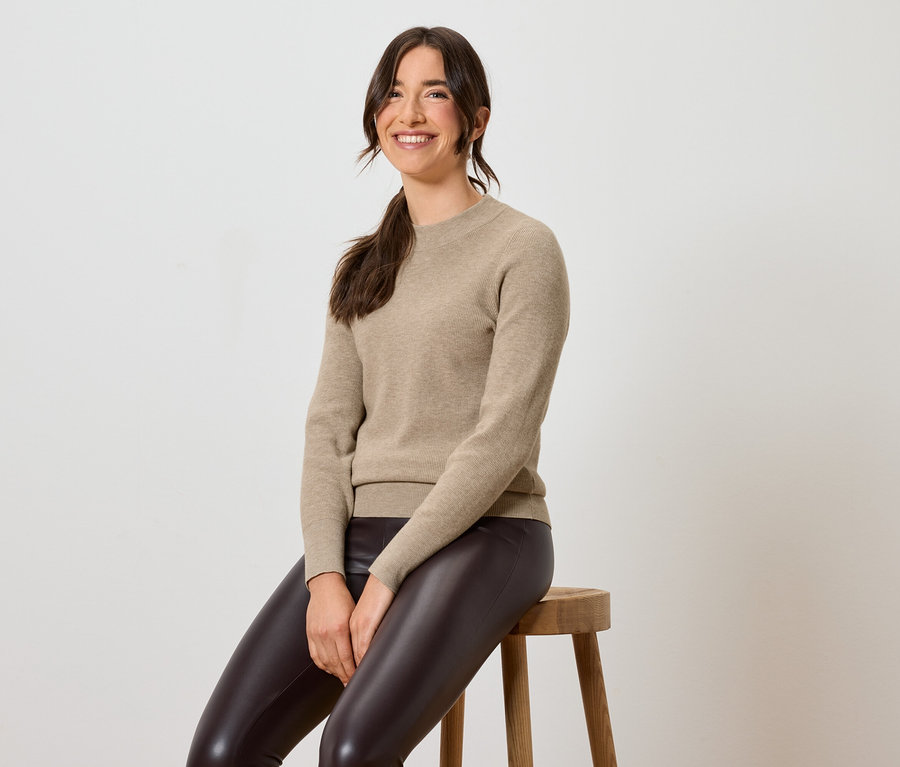 Frau sitzt auf einem Hocker und trägt braune Lederleggings und einen beige Pullover.