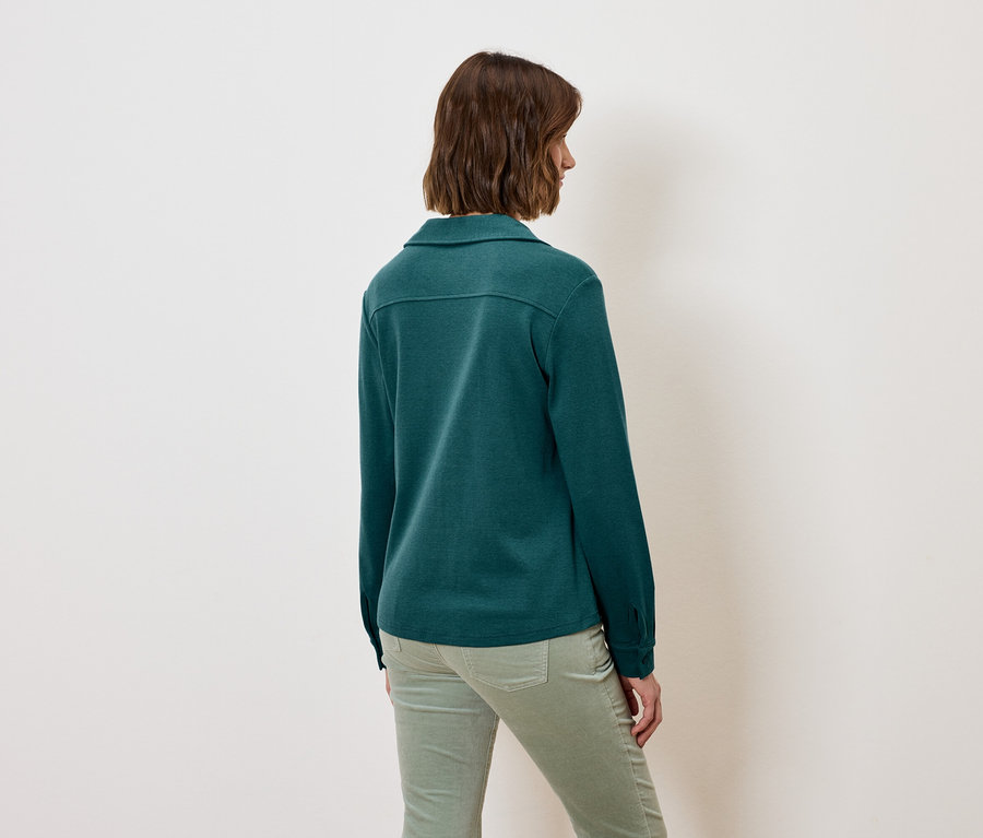 Frau mit dem Rücken zur Kamera trägt ein grünes Langarmhemd und grüne Hosen.