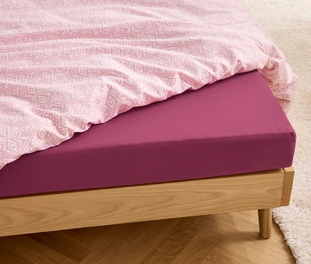 Detailansicht eines Holzbettes mit einem beerenfarbenen Jersey-Spannbettlaken in Normalgröße und rosa Bettwäsche.