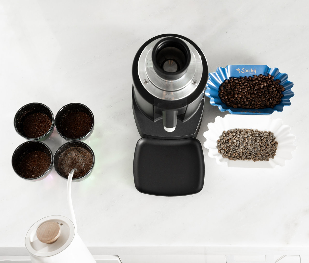Draufsicht auf eine Kaffeemühle, vier Behälter mit Kaffeepulver und zwei Schalen mit Kaffeebohnen auf einer weißen Oberfläche.