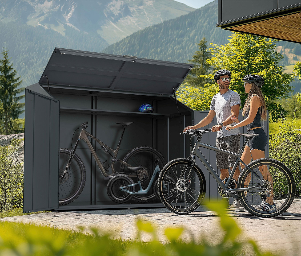 Ein Mann und eine Frau stehen mit Fahrrädern vor einem offenen, grauen Fahrradschuppen in einer bergigen Landschaft.