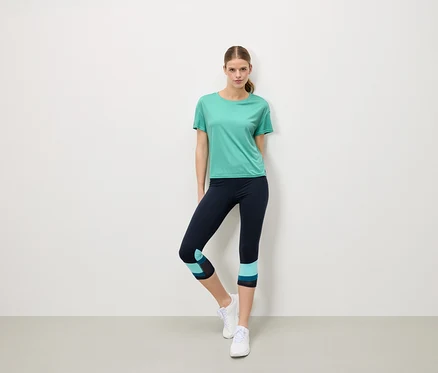 Frau steht vor einer weißen Wand und trägt ein türkisfarbenes T-Shirt, marineblaue Leggings mit türkisfarbenen Details und weiße Turnschuhe.