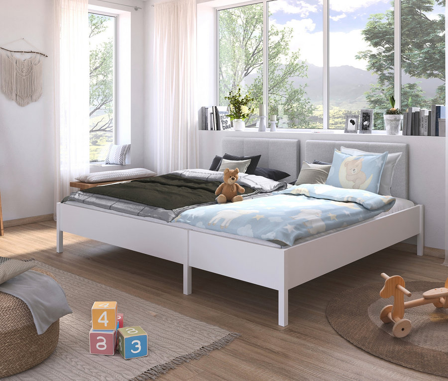 Zwei weiße Betten mit hellgrauen Kopfteilen stehen nebeneinander in einem hellen Raum. Auf dem rechten Bett liegt eine hellblaue Bettdecke mit einem Bärenkissen, auf dem linken eine grüne Decke und ein Teddybär.