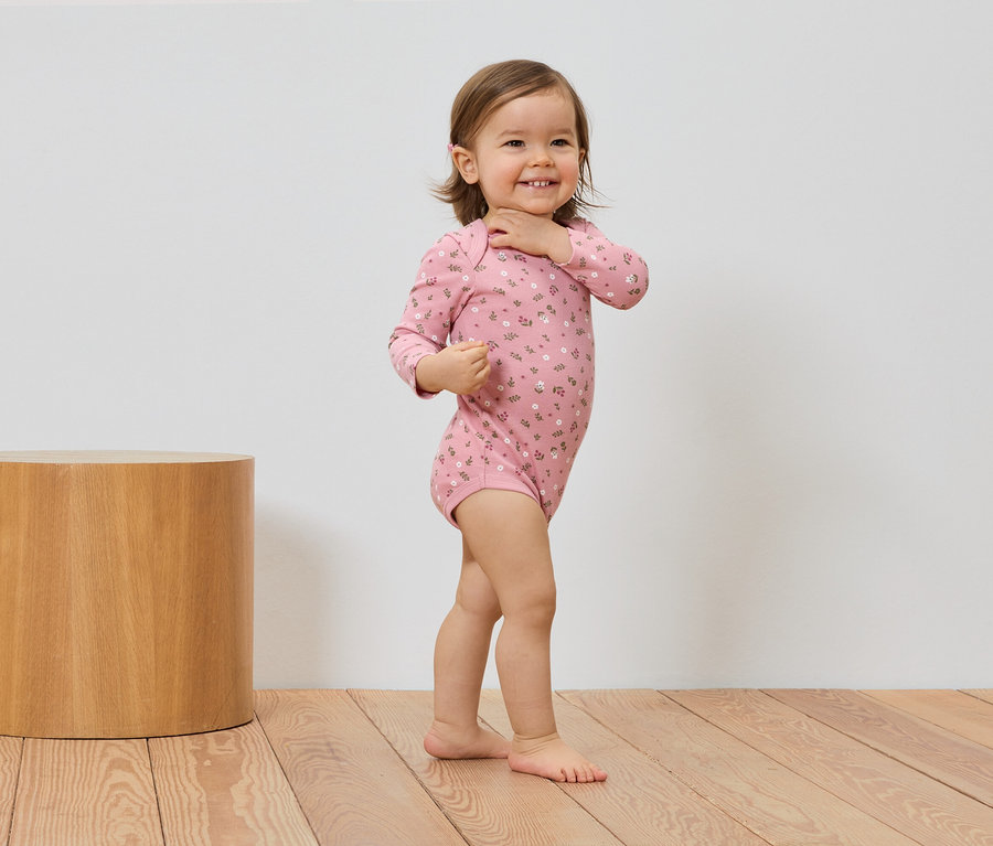 Ein lächelndes Kleinkind in einem rosa Body mit Blumenmuster steht neben einem Holzhocker auf einem Holzboden.