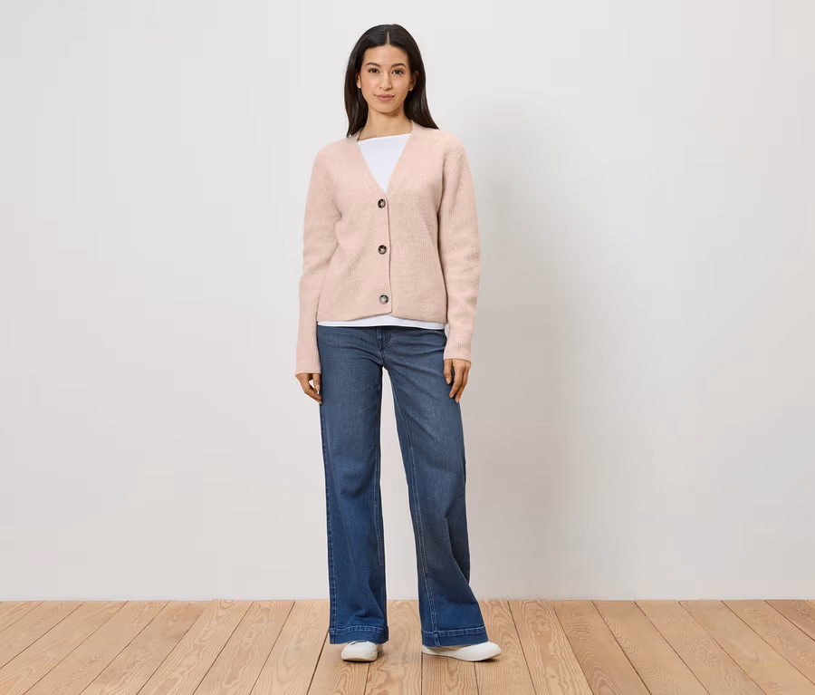 Frau mit rosa Strickjacke, weißem T-Shirt, blauen Jeans und weißen Schuhen steht vor einer weißen Wand.