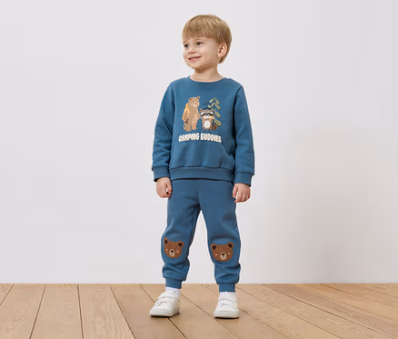 Ein Junge mit blonden Haaren trägt einen blauen Pullover mit Bären- und Waschbär-Motiv und blaue Hosen mit Bärenflicken auf den Knien.