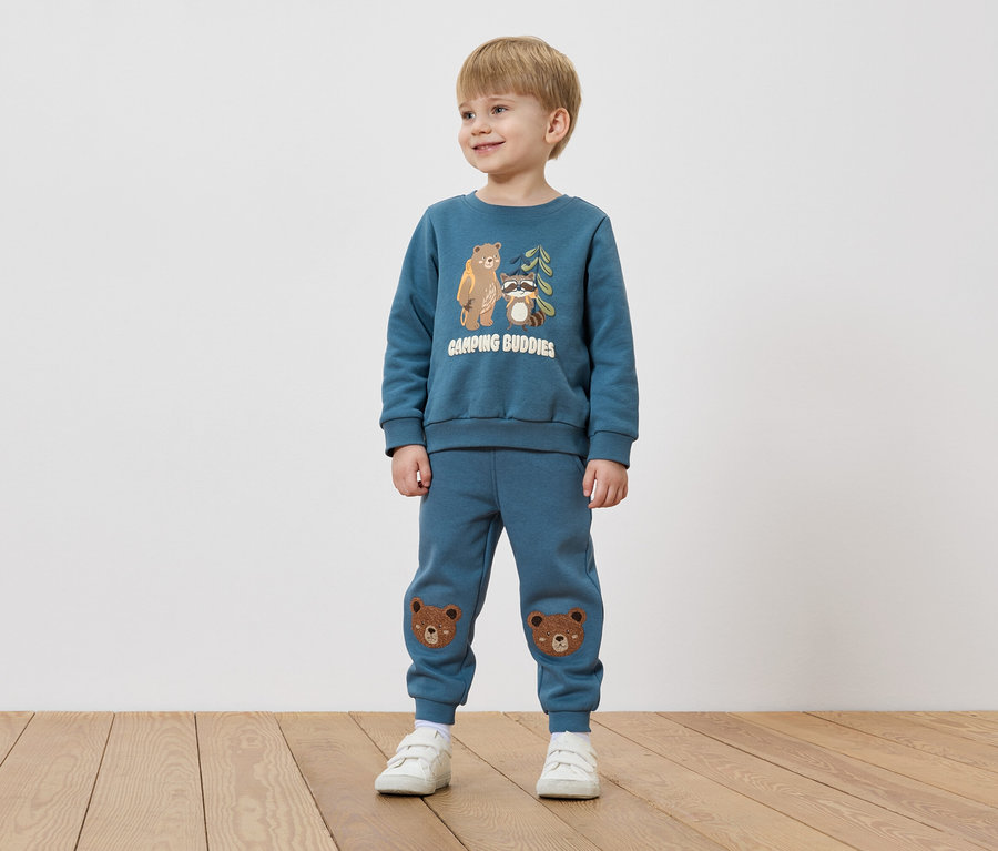 Ein Junge mit blonden Haaren trägt einen blauen Pullover mit Bären- und Waschbär-Motiv und blaue Hosen mit Bärenflicken auf den Knien.
