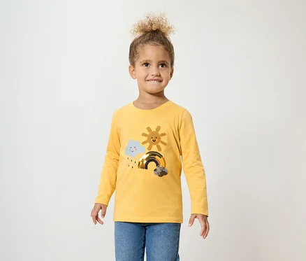 Mädchen mit lockigem Haar trägt ein gelbes Langarmshirt mit einer Wolke, einer Sonne und einem Regenbogen und blaue Jeans.