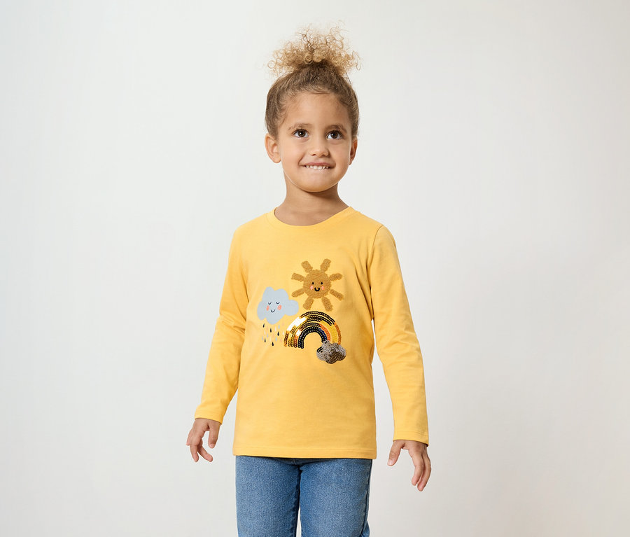 Mädchen mit lockigem Haar trägt ein gelbes Langarmshirt mit einer Wolke, einer Sonne und einem Regenbogen und blaue Jeans.