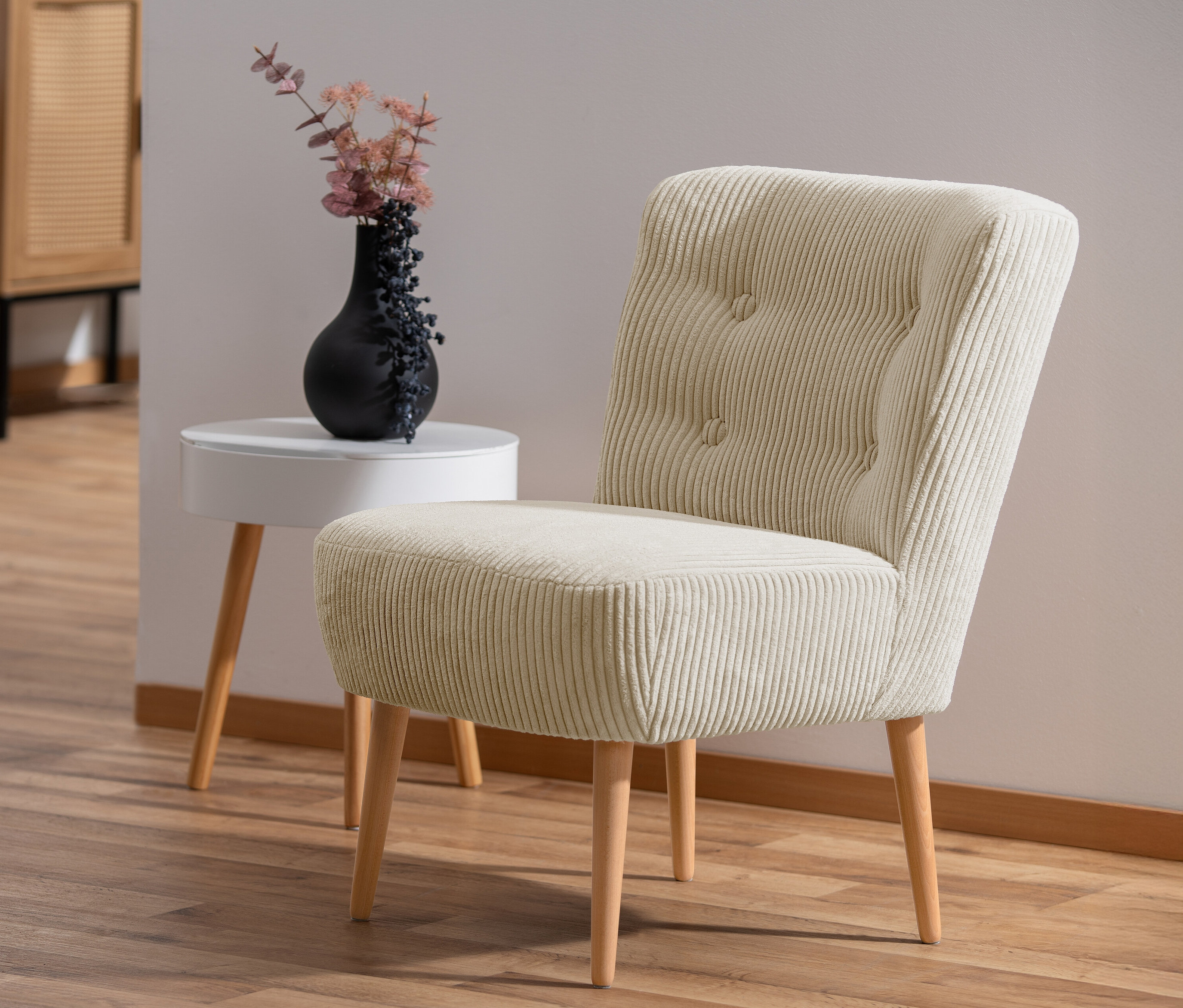 Ein cremefarbener Cocktail-Sessel »Fiona« aus Cordstoff steht auf einem Holzboden. Neben dem Sessel steht ein weißer Beistelltisch mit einer schwarzen Vase.