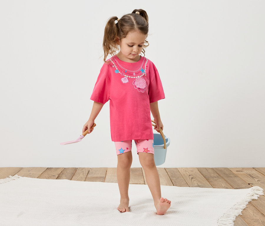 Ein kleines Mädchen mit Zöpfen steht barfuß auf einem weißen Teppich. Sie trägt ein pinkes T-Shirt mit Muscheldekor und rosa Shorts mit Sternen. In ihren Händen hält sie eine Schaufel und einen blauen Eimer.
