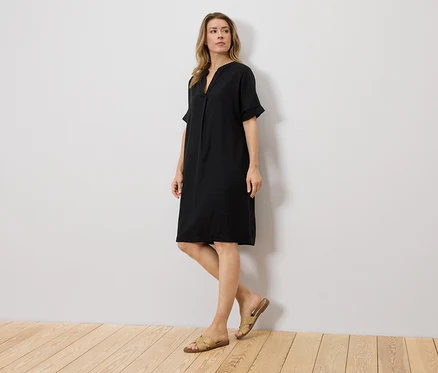 Frau steht vor einer Wand und trägt ein schwarzes Kleid und Sandalen.