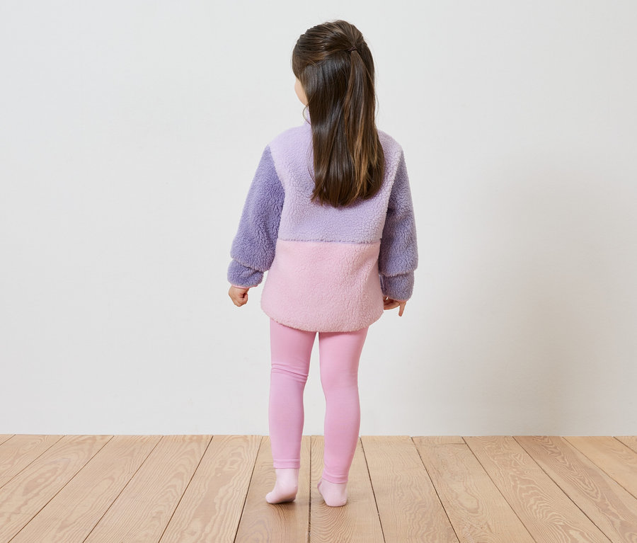 Rückansicht eines Mädchens mit langen, braunen Haaren, das einen lavendelfarbenen und rosa farbblockierten Teddy-Pullover und rosa Leggings trägt.